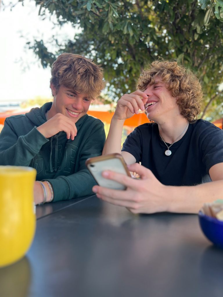 Two teenage boys sitting at a table and laughing while looking at something on a phone