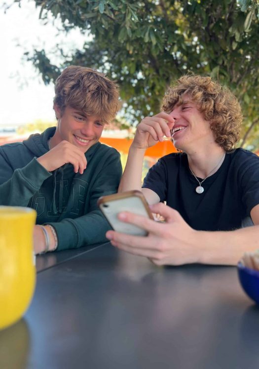 Two teenage boys sitting at a table and laughing while looking at something on a phone